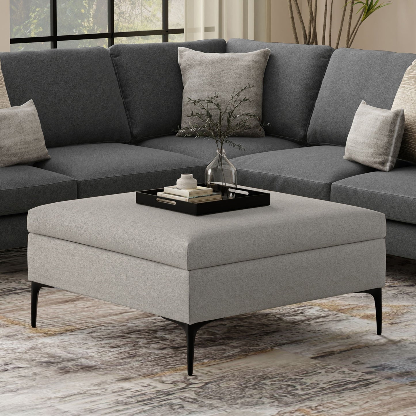 The Rebecca Multifunctional Coffee Table Storage Ottoman with Lift Up Lid in gray sits before a dark gray sectional sofa. A black tray on the ottoman holds a glass vase with greenery and books, adding modern, cozy style to the room.