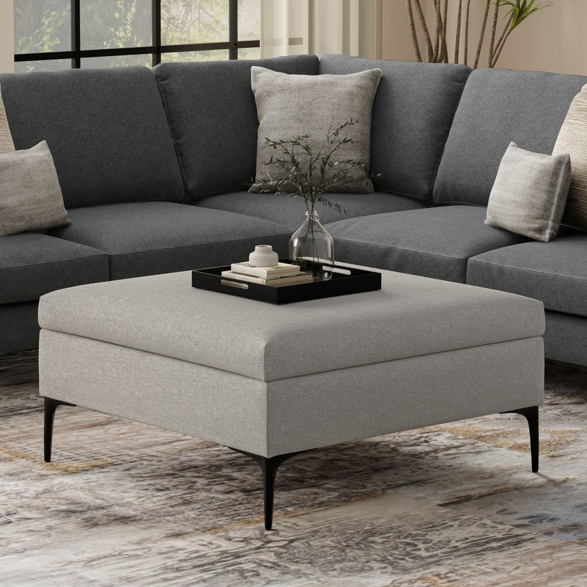 The Rebecca Multifunctional Coffee Table Storage Ottoman with Lift Up Lid in gray sits before a dark gray sectional sofa. A black tray on the ottoman holds a glass vase with greenery and books, adding modern, cozy style to the room.