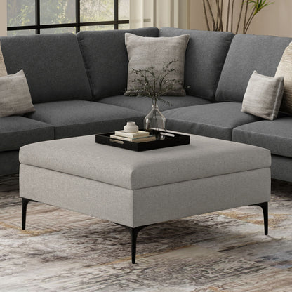 The Rebecca Multifunctional Coffee Table Storage Ottoman with Lift Up Lid in gray sits before a dark gray sectional sofa. A black tray on the ottoman holds a glass vase with greenery and books, adding modern, cozy style to the room.