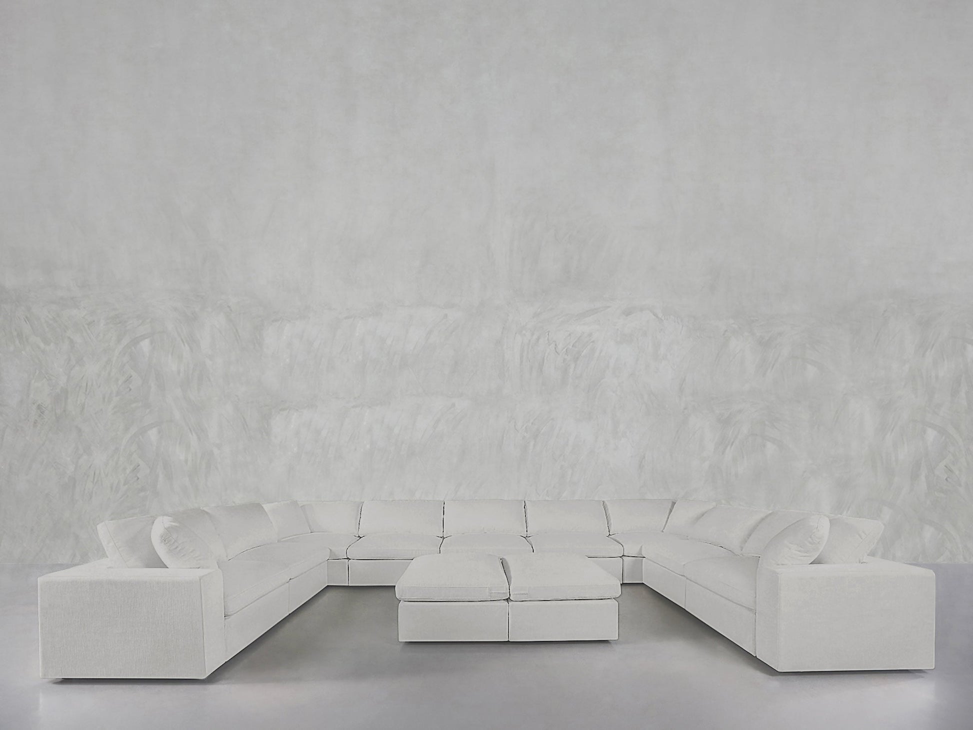 The minimalist, modern space features a 9-Seat Modular U-Sectional with Double Ottoman in white, set against a light gray textured wall and smooth gray floor, emphasizing clean lines and open space.