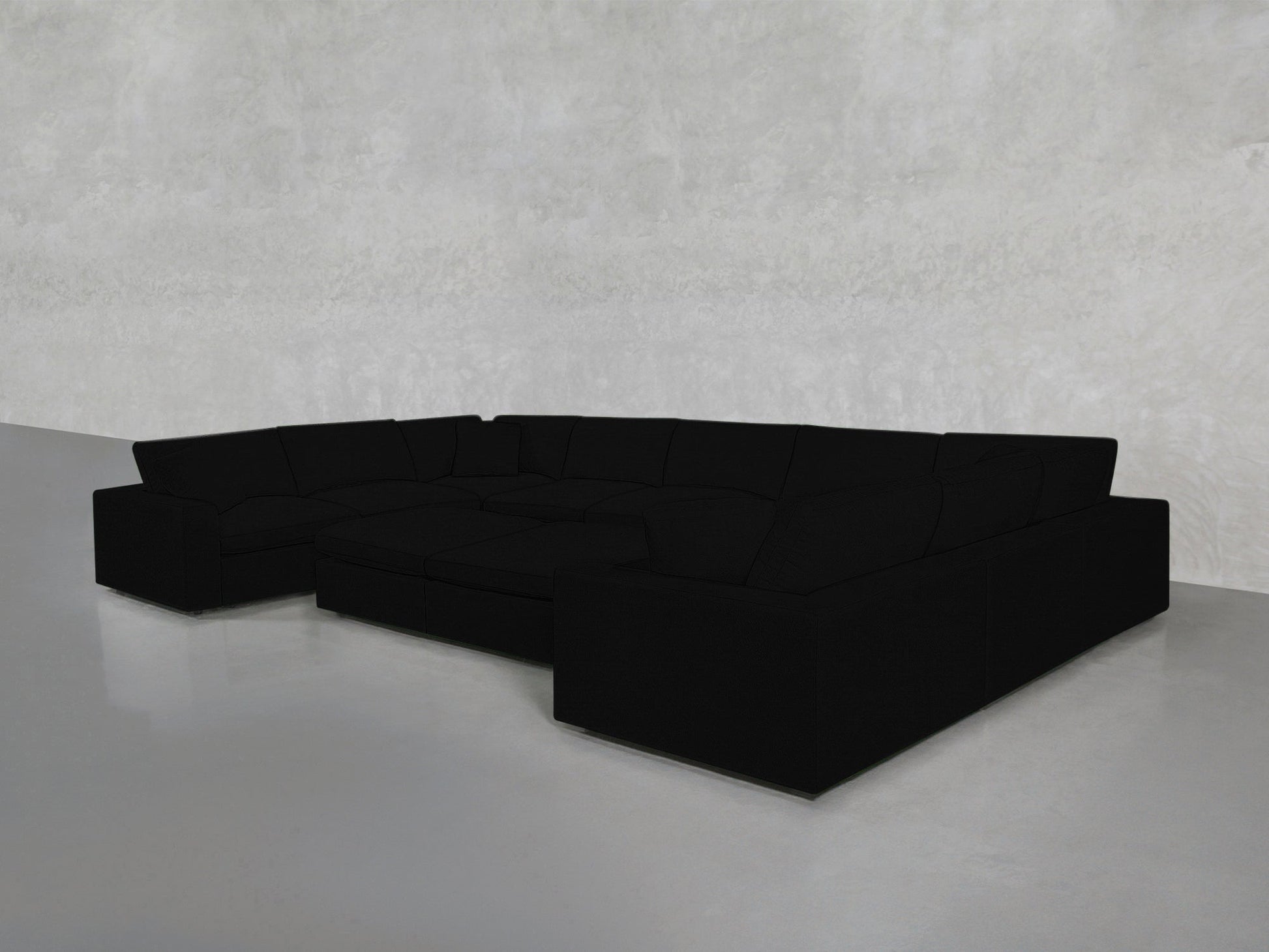 A 9-Seat Modular U-Sectional with Double Ottoman in black, featuring deep seats and clean lines, sits in an empty room with a smooth light gray concrete floor and a textured light gray wall.