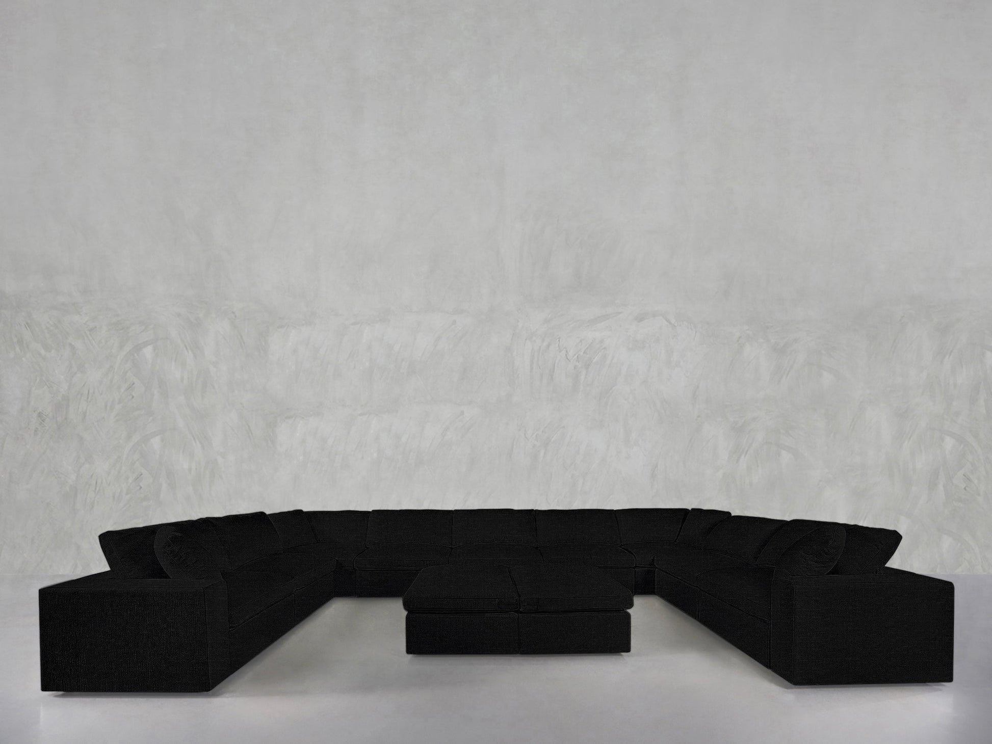 A 9-Seat Modular U-Sectional with Double Ottoman in black stands against a light gray textured wall in a minimalistic room featuring a white floor.