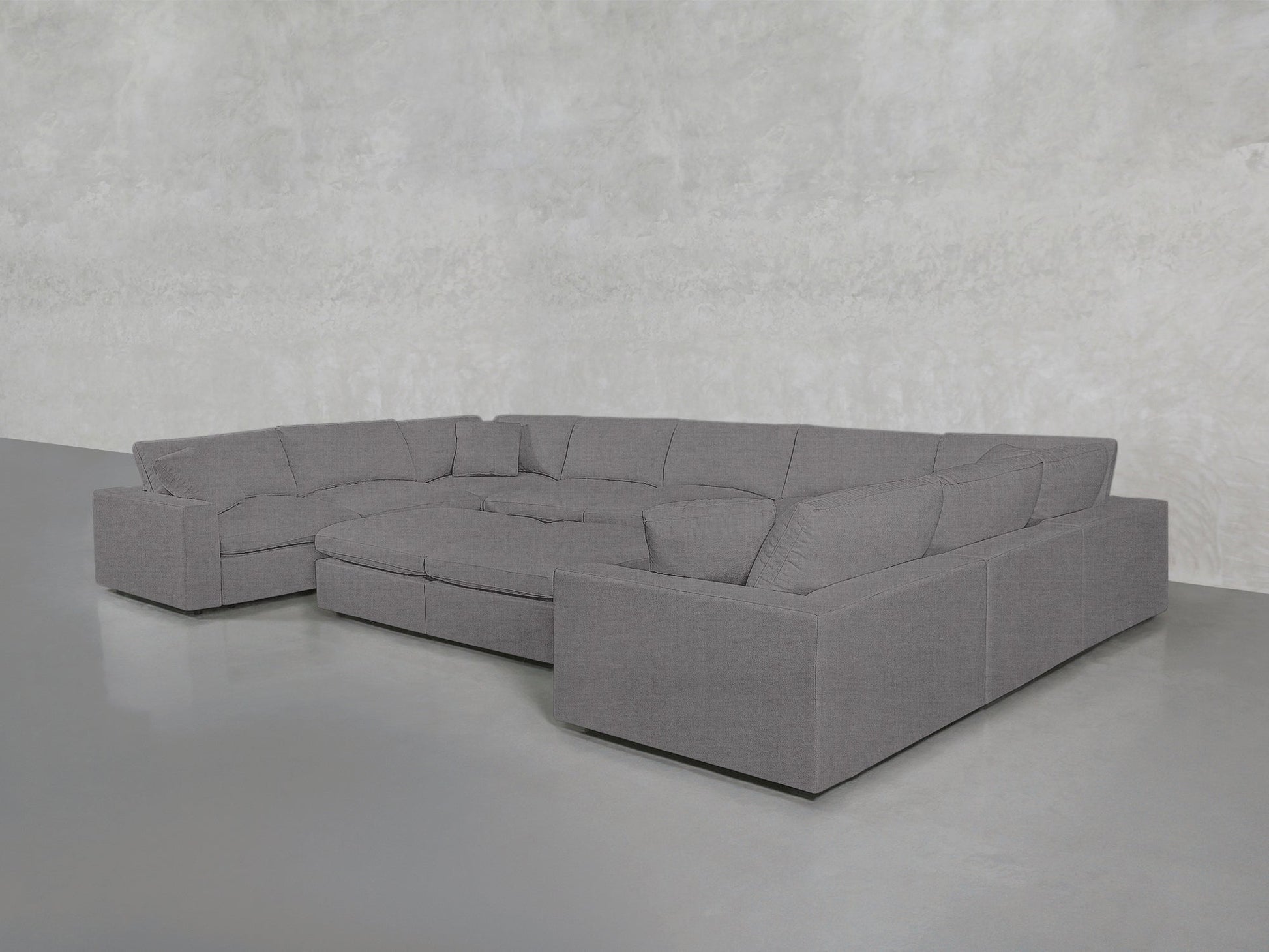 A spacious, modern 9-Seat Modular U-Sectional with Double Ottoman in gray is set in an empty room featuring a light gray textured wall and a smooth gray floor.