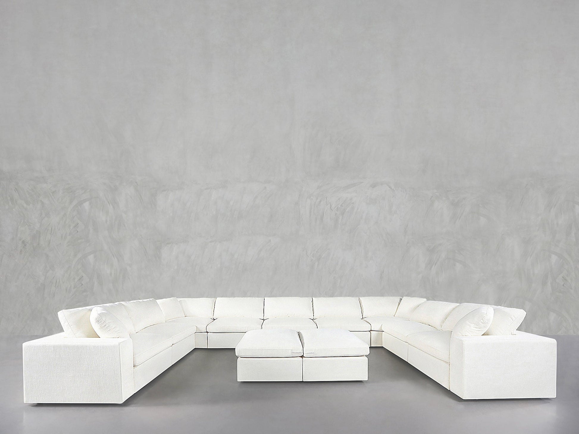 A 9-Seat Modular U-Sectional with Double Ottoman in white stands against a light gray textured wall and gray floor, creating a minimalist, modern look.