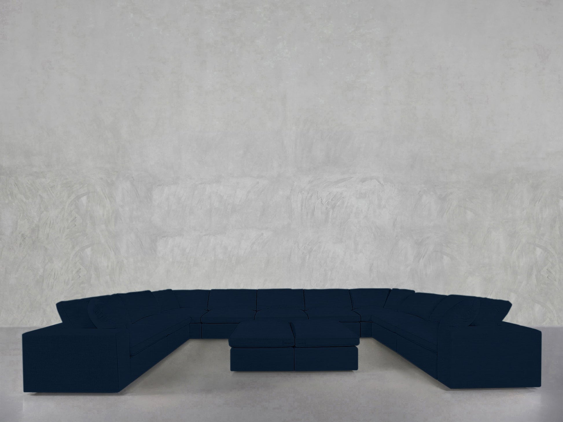 A 9-Seat Modular U-Sectional with Double Ottoman in dark blue is placed against a plain, light gray textured wall and floor.