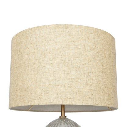 A close-up of the Aurora table lamp features a large, beige linen cylindrical shade with textured fabric, resting on a ribbed clear base. The metal rod beneath is gold-toned, set against a plain white background.