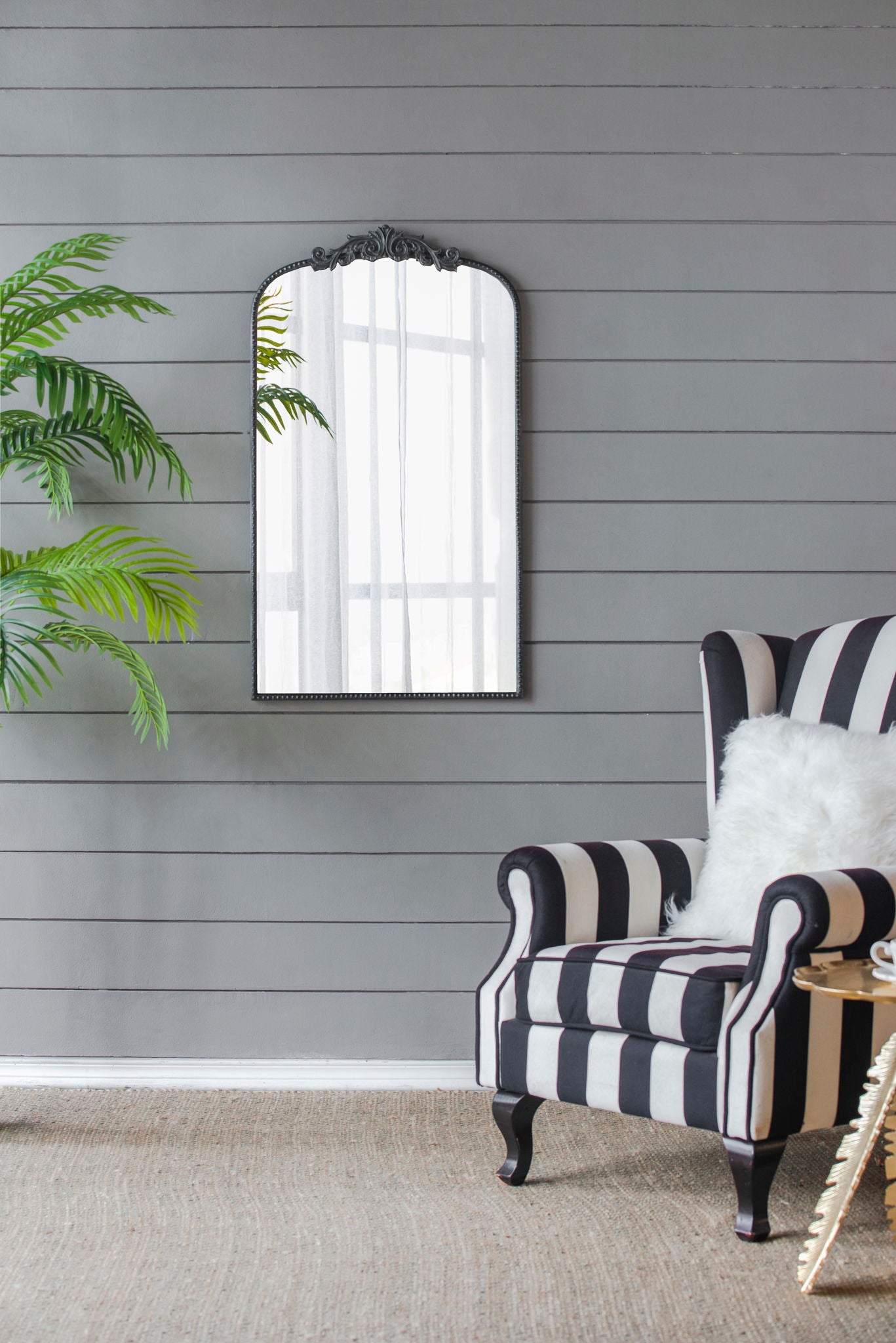 A black and white striped armchair with a white cushion sits by a round side table; a potted palm and the Arch Mirror, Baroque Inspired Wall Decor, are against a gray shiplap wall with natural light reflecting in the mirror.