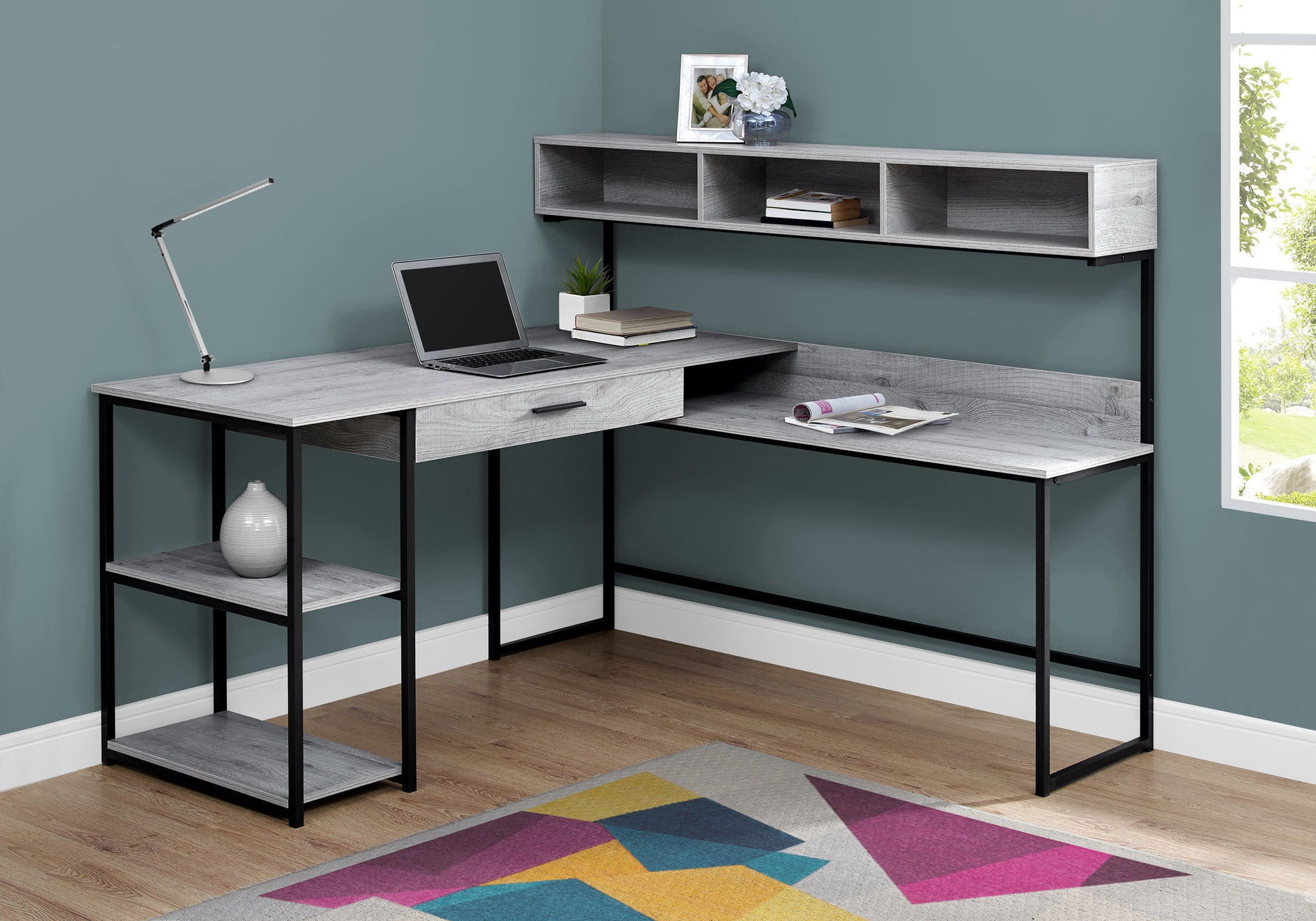 A contemporary L-shaped Computer Desk with storage drawers for home office use holds a laptop and decor, positioned in a corner room with teal walls, a window, and a colorful geometric rug on wood flooring.