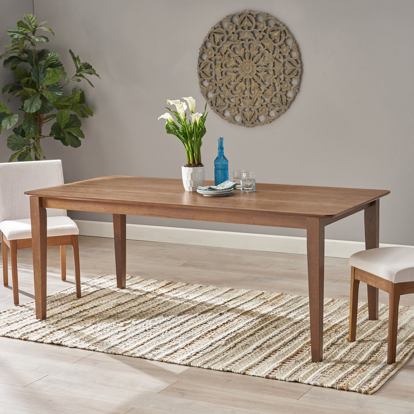 The Farmhouse Charm Dining Table With Square Leg is paired with two upholstered chairs on a striped rug, topped with a vase of white flowers, a blue bottle, and two glasses. A decorative wall piece and potted plant complete the background.