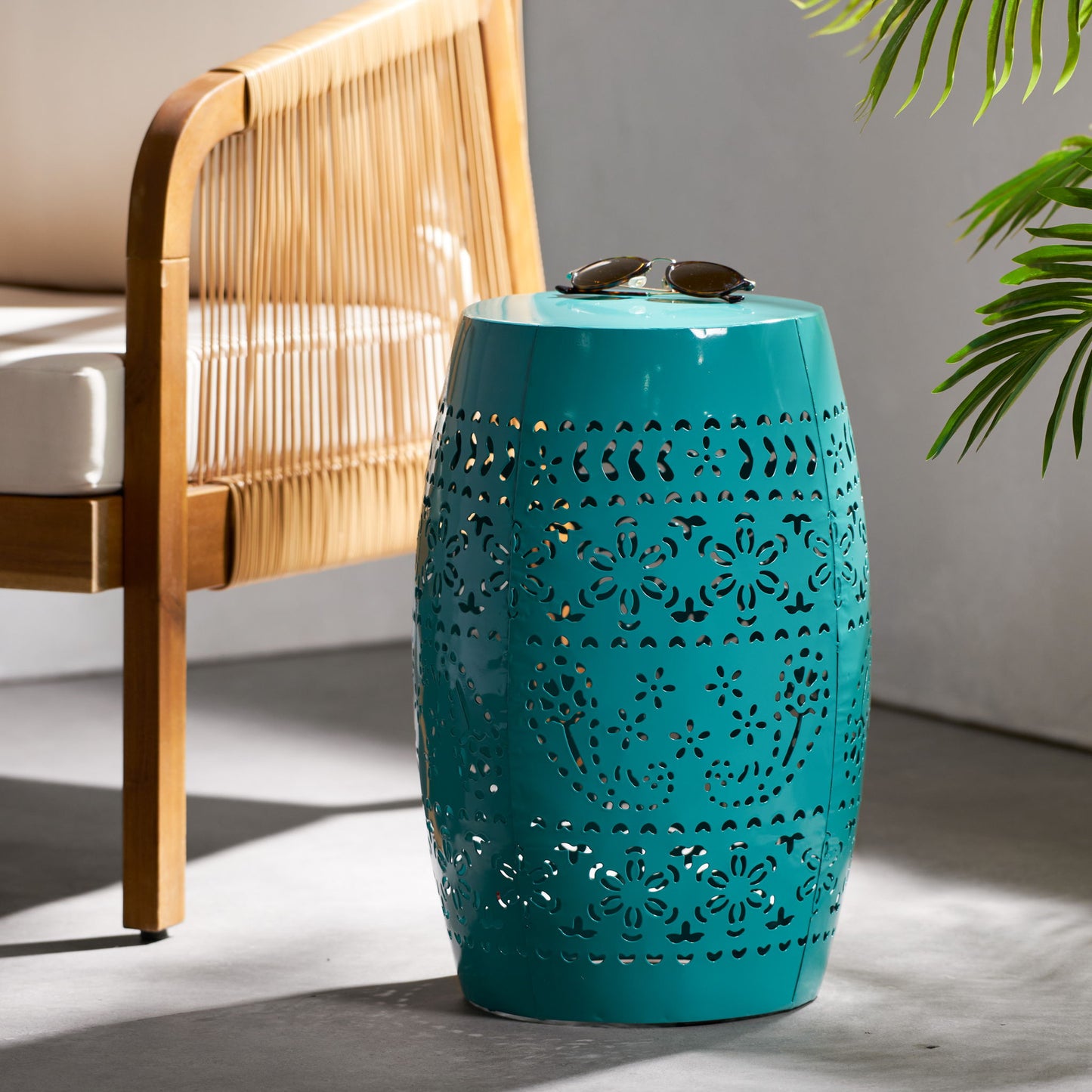 A 12" Outdoor Lace Cut Side Table with a turquoise ceramic finish stands beside a wooden chair with rope details. Sunglasses rest atop the table, and a green plant appears in the background.