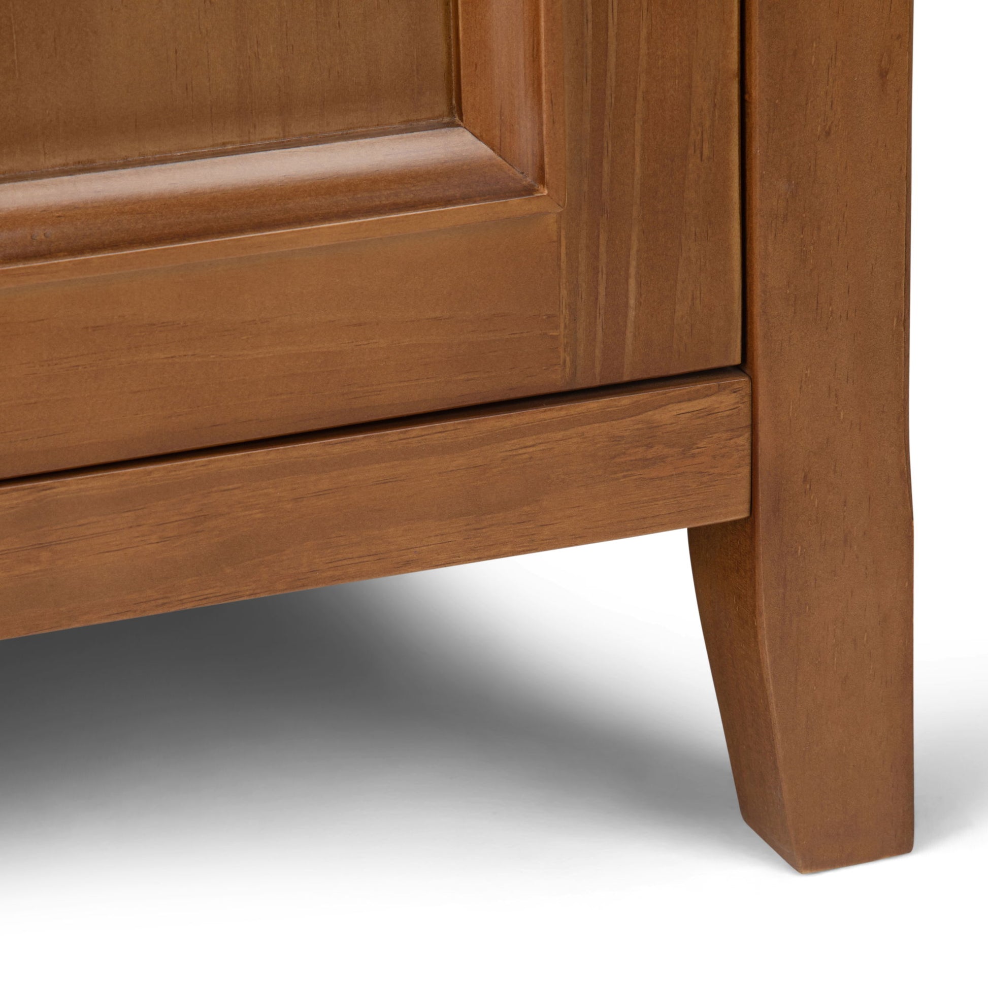 Close-up of the Amherst - Handcrafted Medium Storage Cabinet's bottom corner, highlighting a leg and panel with a warm brown finish. The cabinet rests on a white surface.