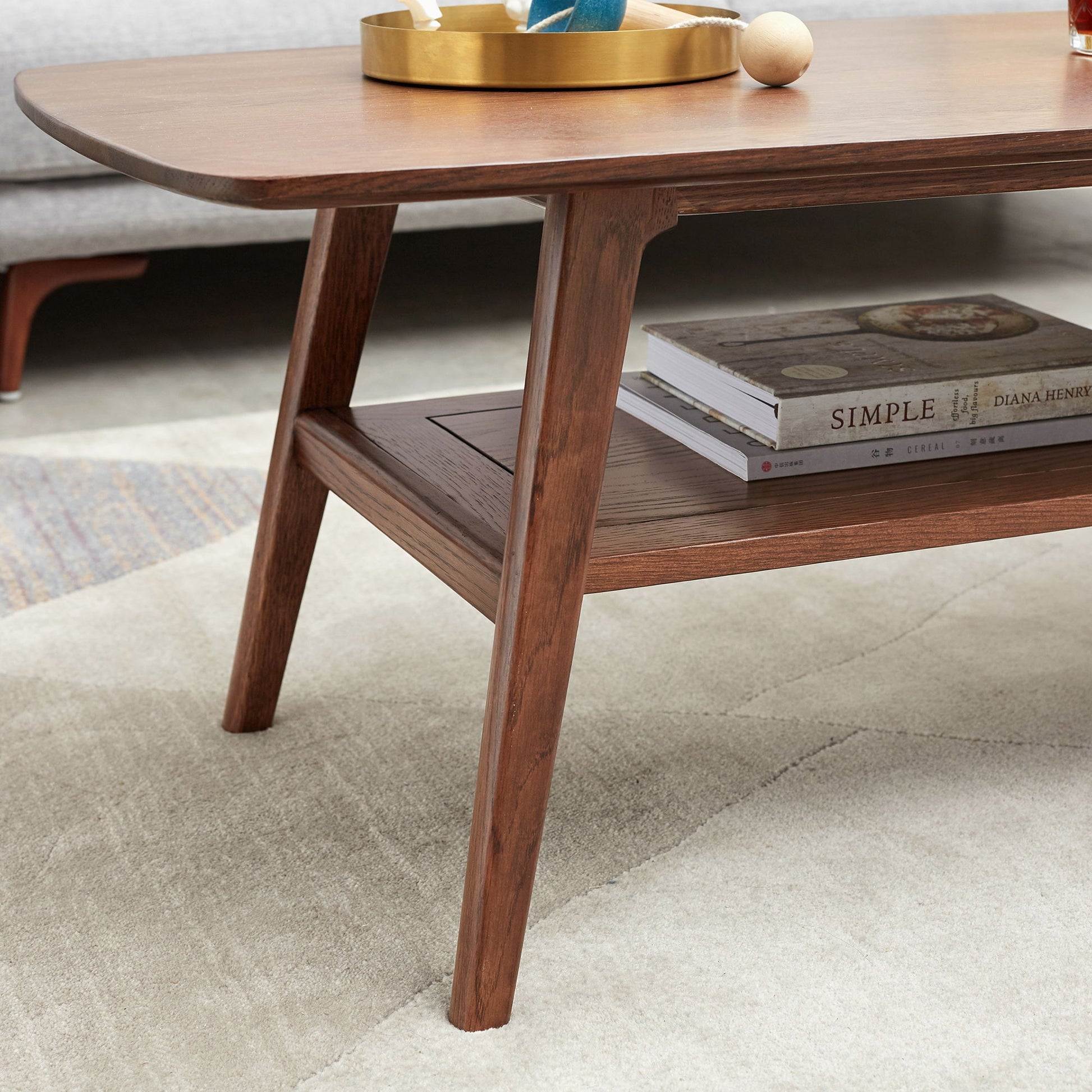 A close-up of the Low Coffee Table With Storage Shelf featuring tapered legs, a gold tray with décor on top, and two hardcover books on the lower shelf. The table rests on a light patterned rug.