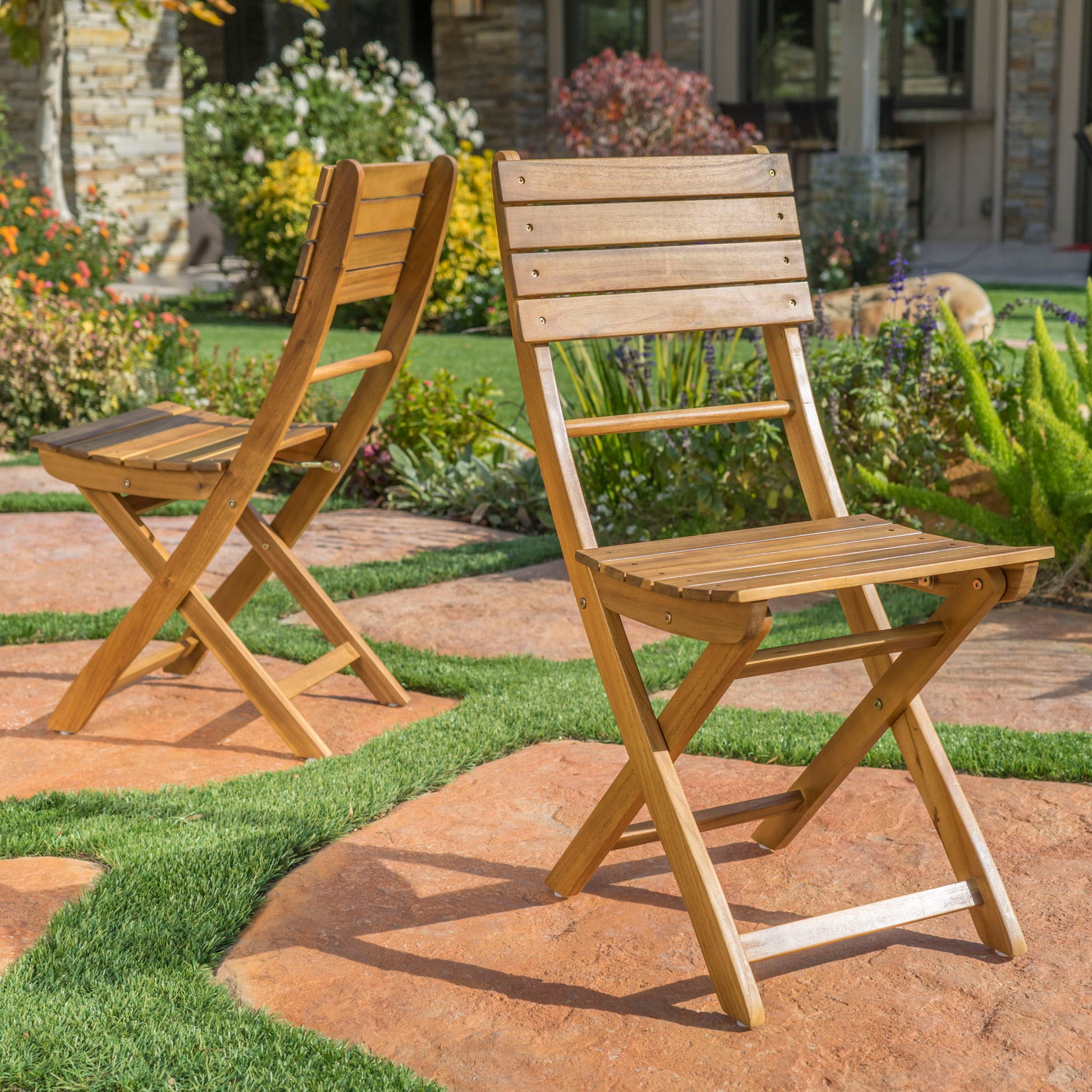 Two Outdoor Foldable Wood Dining Chairs With Slat Design (Set of 2) are set on a stone and grass patio in a garden, surrounded by flowers and greenery under bright sunlight.