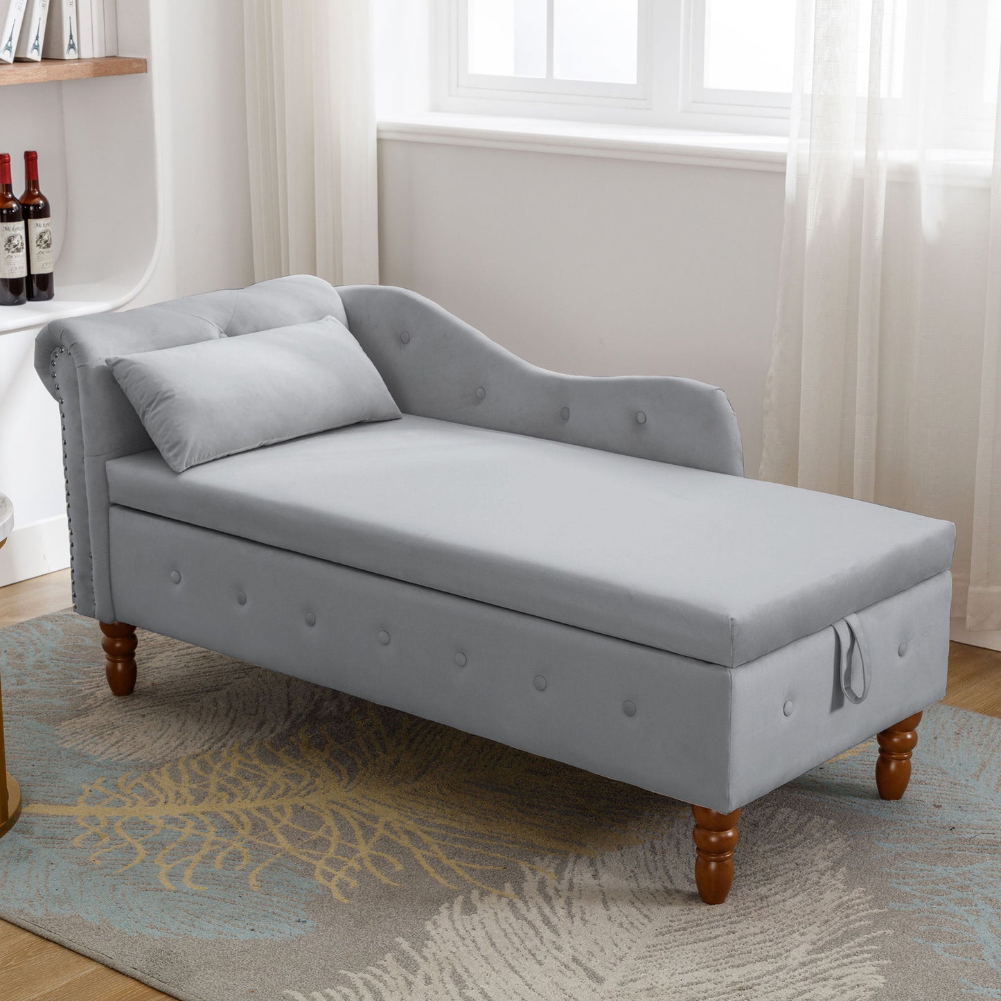 The Chaise Lounge Indoor, a modern velvet lounge chair with storage, pillow, wooden legs, and nailhead trim, sits on a patterned rug near a window with sheer curtains in a bright, cozy room.