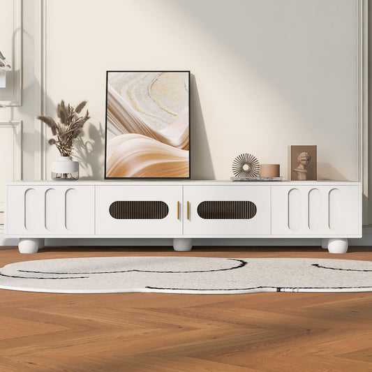 A Modern TV Stand for TVs up to 80" with glass door, 2 drawers, cabinets, solid wood legs, and metal handles displays decor like framed art, a vase with dried plants, a sculpture, and an ornament. A white rug sits on the wooden floor.