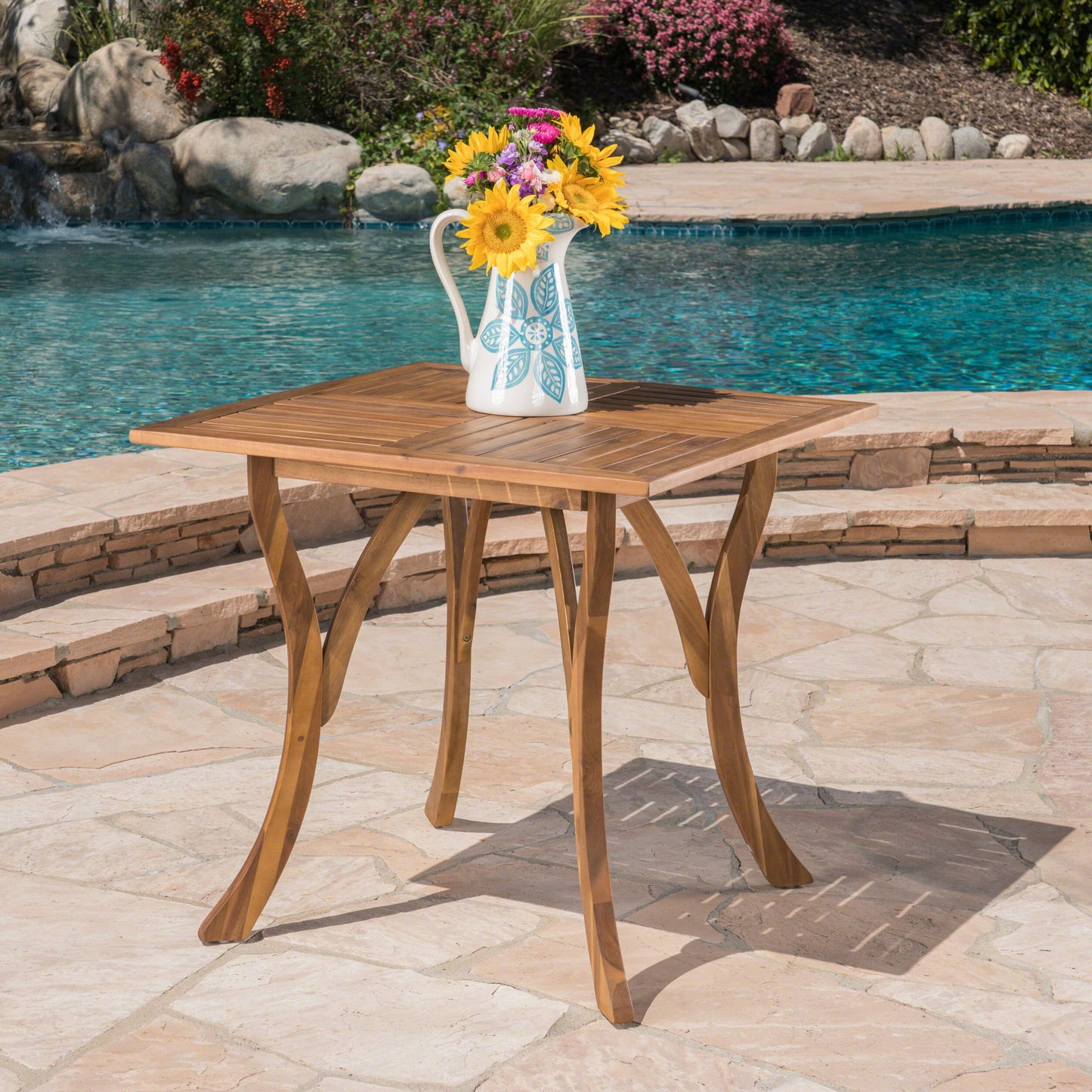 An Outdoor Rectangular Table With Curved Legs And Perpendicular Wooden Slats Table Top stands on a stone patio by the pool, holding a white pitcher with colorful flowers, with shrubs and rocks in the background.