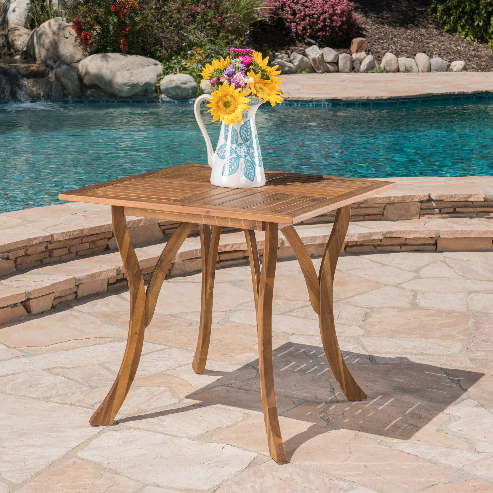 An Outdoor Rectangular Table With Curved Legs And Perpendicular Wooden Slats Table Top stands on a stone patio by the pool, holding a white pitcher with colorful flowers, with shrubs and rocks in the background.