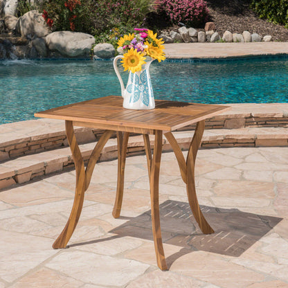 An Outdoor Rectangular Table With Curved Legs And Perpendicular Wooden Slats Table Top stands on a stone patio by the pool, holding a white pitcher with colorful flowers, with shrubs and rocks in the background.
