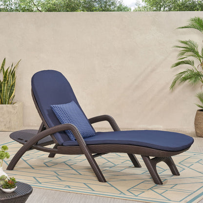 The Mikael - Chaise Lounge With Cushion, featuring a navy blue cushion and patterned pillow, sits on a patterned rug among potted plants against a beige wall in a garden setting.