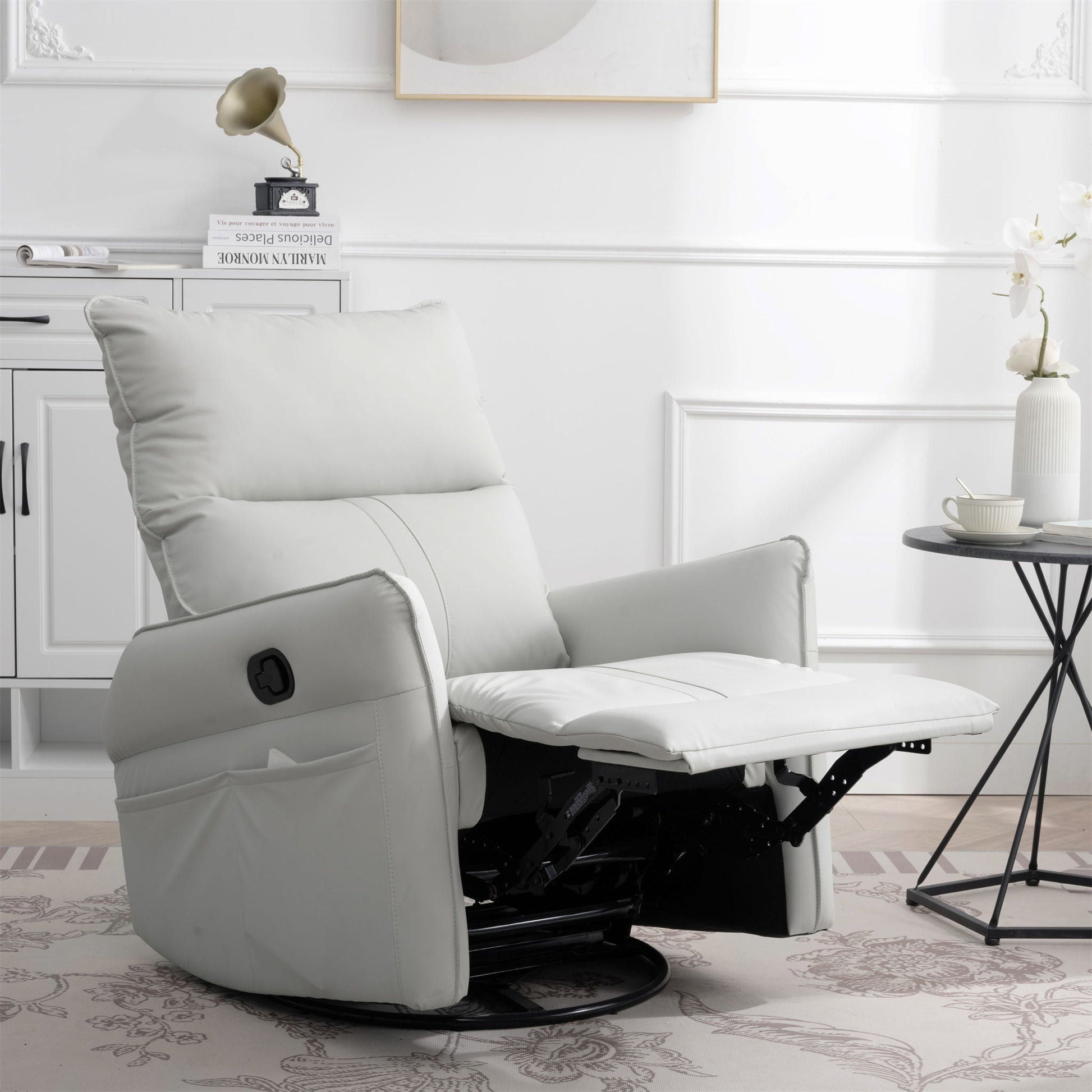 The 360° Modern Small Rocking Swivel Recliner Chair with Side Pocket, in white, features an extended footrest and sits in a bright living room with white walls, a round side table, teacup, and decorative flower vase nearby.