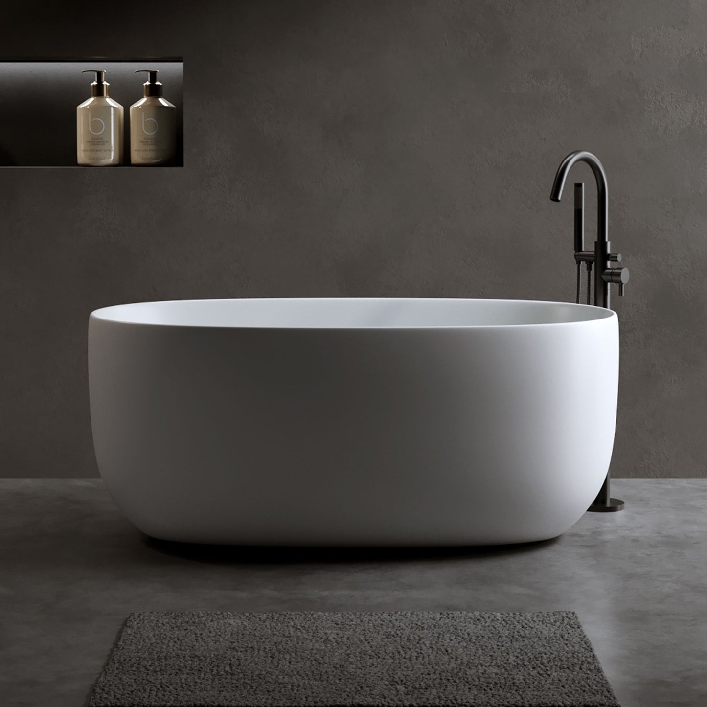 A matte white Independent Solid Surface Resin Stone Bathtub with pop-up drainage and overflow pipes stands on a gray floor. Two bottles rest in a recessed shelf on the dark gray wall, with a textured gray bath mat in front of the tub.