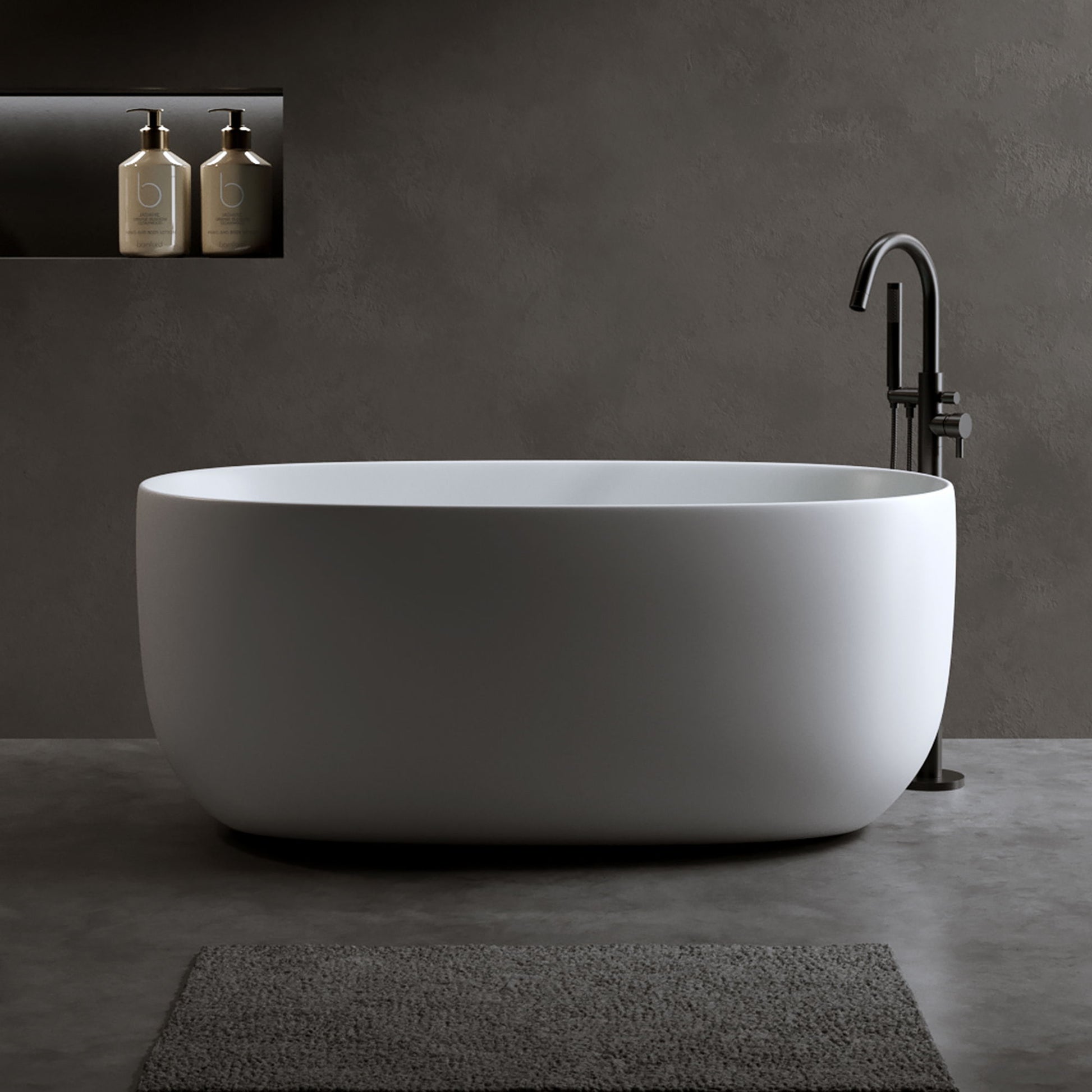 A matte white Independent Solid Surface Resin Stone Bathtub with pop-up drainage and overflow pipes stands on a gray floor. Two bottles rest in a recessed shelf on the dark gray wall, with a textured gray bath mat in front of the tub.