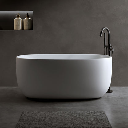 A matte white Independent Solid Surface Resin Stone Bathtub with pop-up drainage and overflow pipes stands on a gray floor. Two bottles rest in a recessed shelf on the dark gray wall, with a textured gray bath mat in front of the tub.