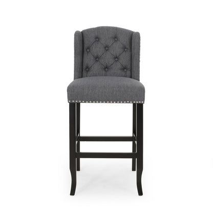 The Vienna Contemporary Fabric Tufted Wingback Counter Stools (Set of 2) features gray upholstery, tufted backrest, silver nailhead trim, and dark wooden legs, shown from the front on a plain white background.