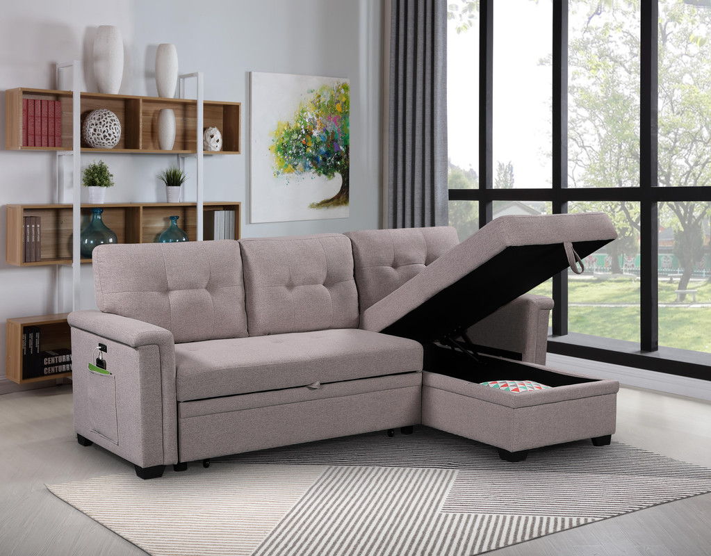 A modern living room with the Ashlyn Reversible Sleeper Sectional Sofa featuring storage chaise, USB ports, and pocket, set on a patterned rug near large windows. Background includes shelves, books, decor, and a colorful tree painting.