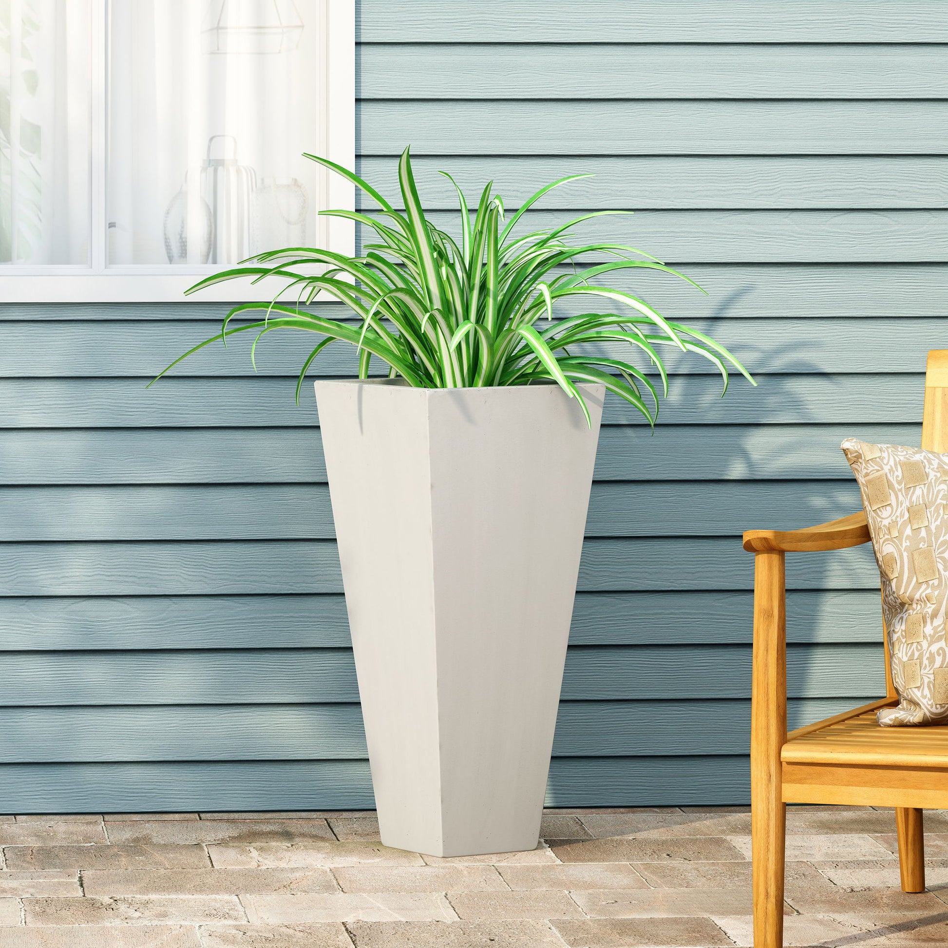 An Outdoor Modern Cast Stone Planter with a spider plant sits on a stone patio before blue siding, beside a wooden chair with a patterned cushion.