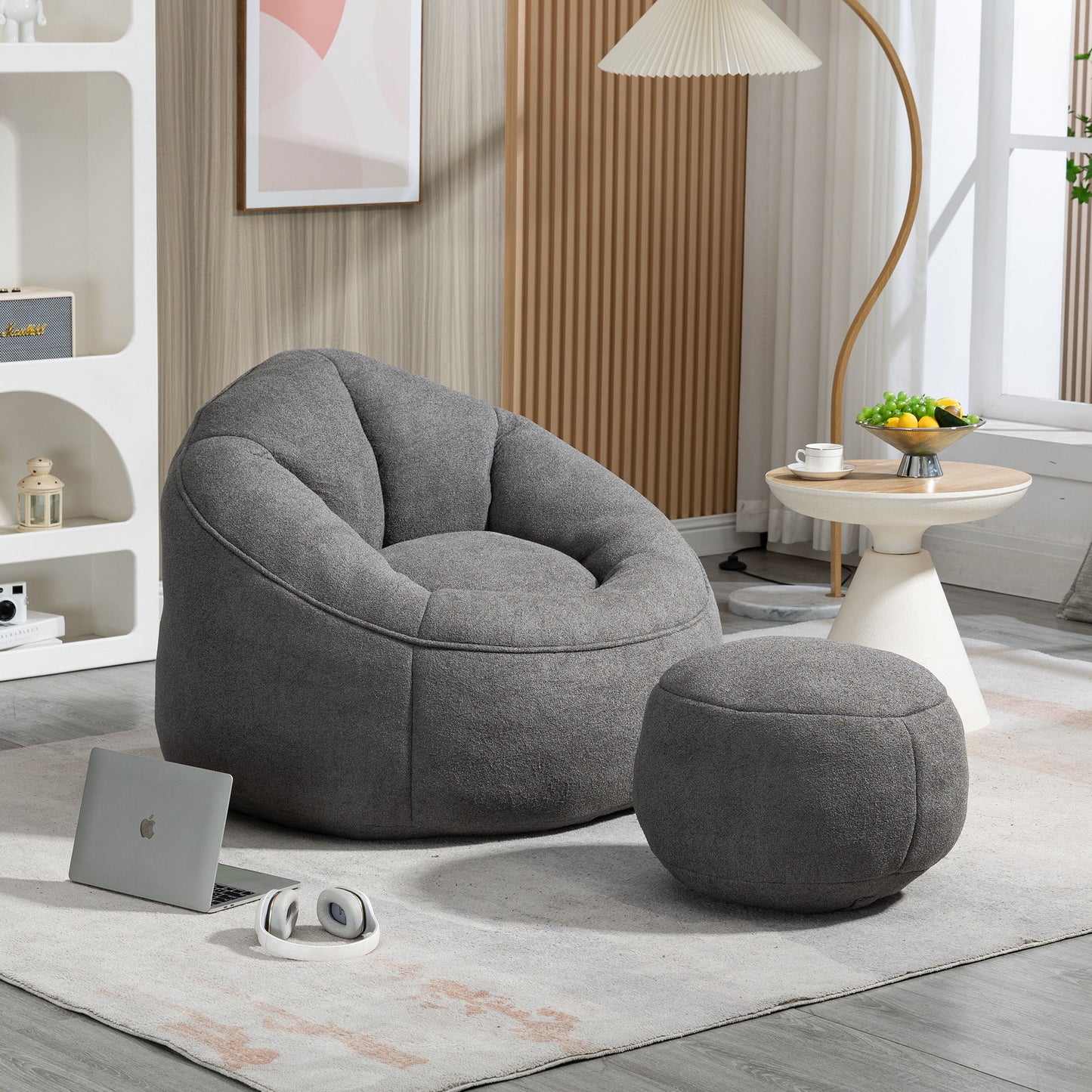 A cozy living room featuring the Bedding Bean Bag Sofa Chair High Pressure Foam with Footrest on a light rug, beside a round table with fruit, a white lamp, a laptop, headphones on the floor, and shelving in the background.