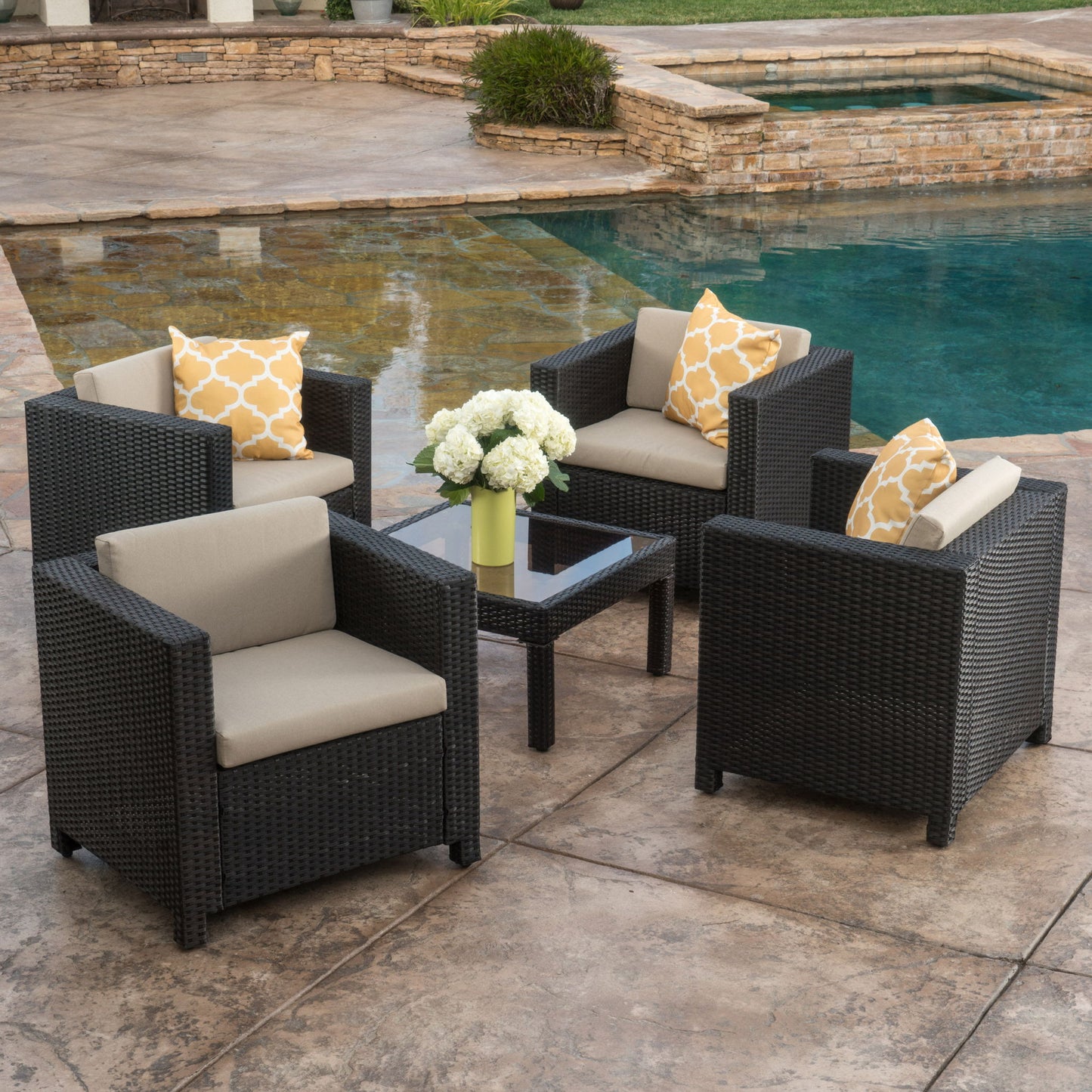 Four Stylish Outdoor Club Chairs with weather-resistant beige cushions and yellow-patterned pillows surround a glass coffee table topped with a vase of white flowers, all arranged by a pool and stone patio in the backyard.