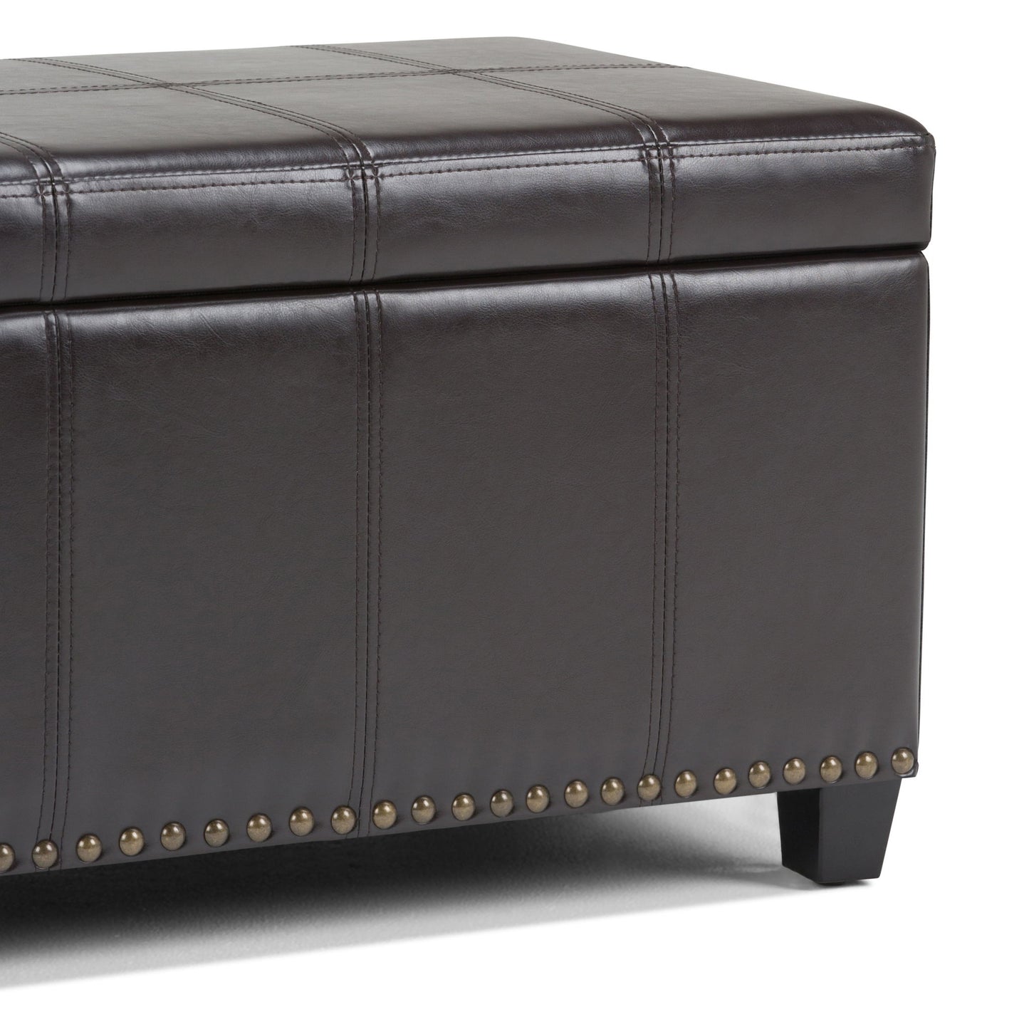 A close-up of the Amelia - Transitional Storage Ottoman Bench in dark brown leather, featuring stitched panels, a lift-top lid for storage, brass nailhead trim at the base, and short black legs.