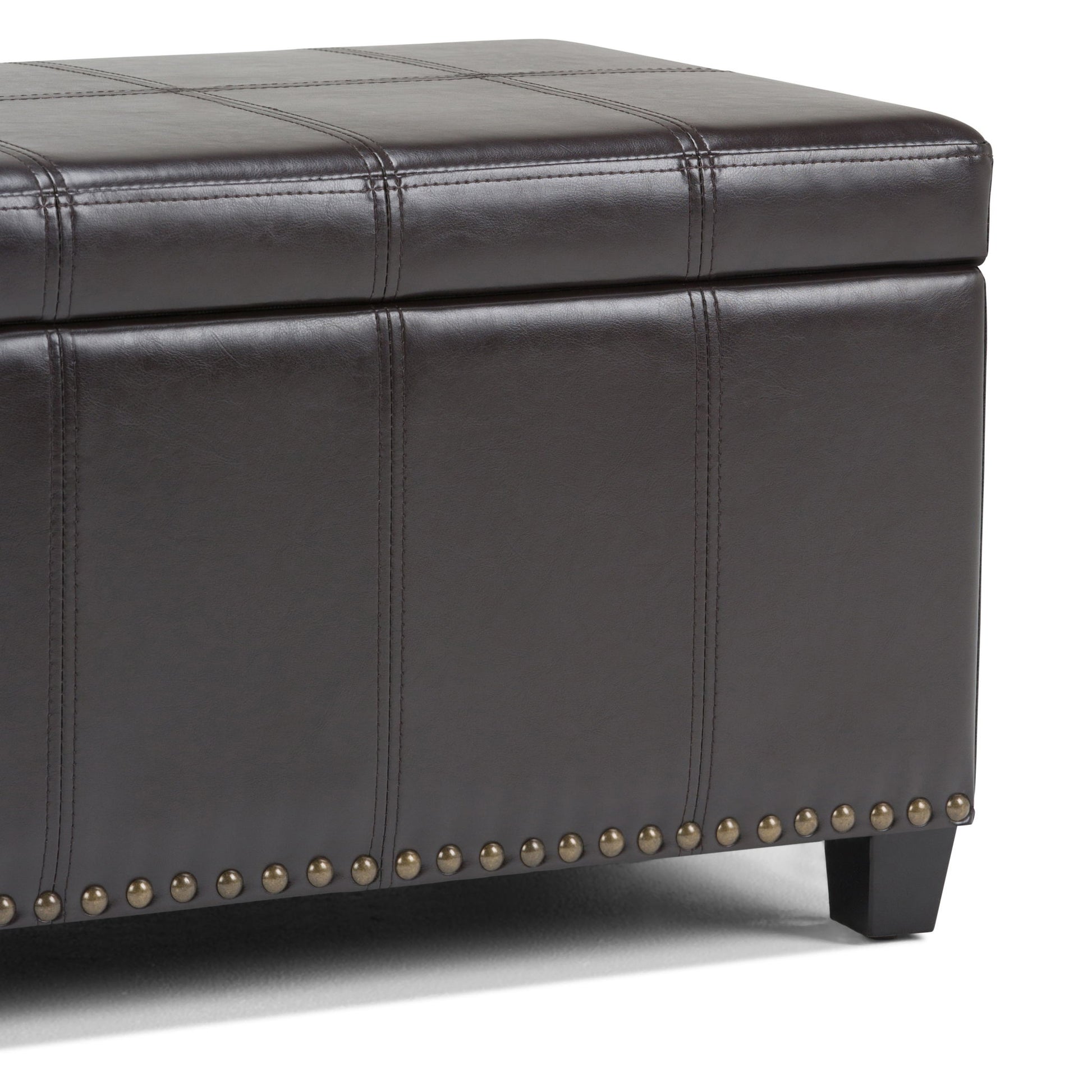A close-up of the Amelia - Transitional Storage Ottoman Bench in dark brown leather, featuring stitched panels, a lift-top lid for storage, brass nailhead trim at the base, and short black legs.