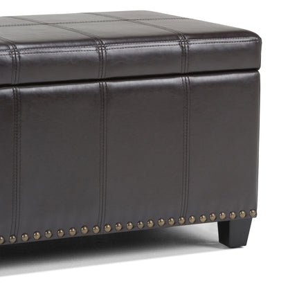 A close-up of the Amelia - Transitional Storage Ottoman Bench in dark brown leather, featuring stitched panels, a lift-top lid for storage, brass nailhead trim at the base, and short black legs.