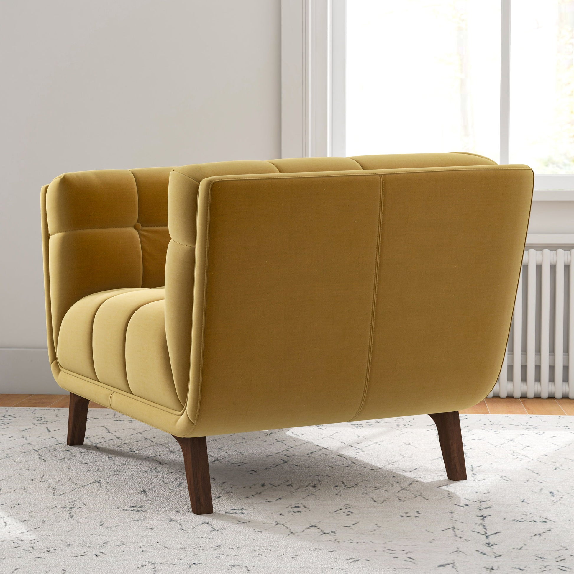 The Addison Mid-Century Modern Lounge Chair in mustard yellow with tufted upholstery and wooden legs sits on a light-patterned rug in a bright room near a window and radiator.