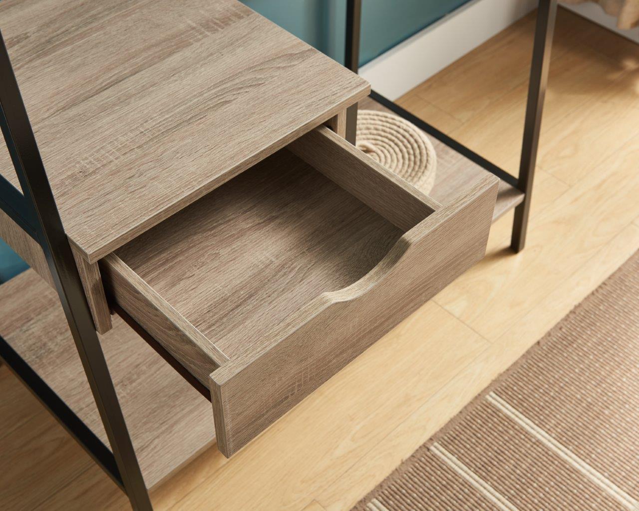 A close-up of the Metal Clothes Rack Open Wardrobe, featuring a wooden drawer, black metal frame, and a nearby woven mat on the wooden floor. Part of a beige rug with white stripes is visible.