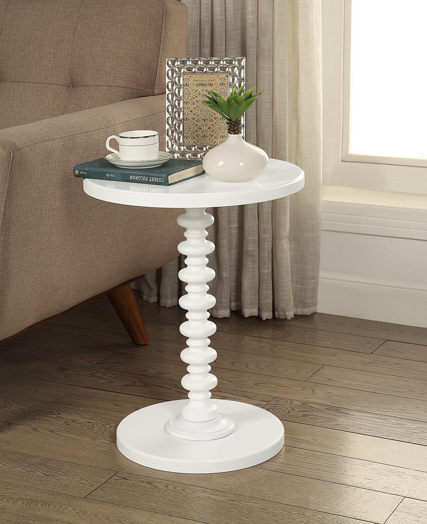The Acton - Accent Table - White, featuring a round top and sculpted pedestal base, stands on a wood floor beside a beige sofa. On it are two books, a white mug, a framed photo, and a small white vase with green leaves.