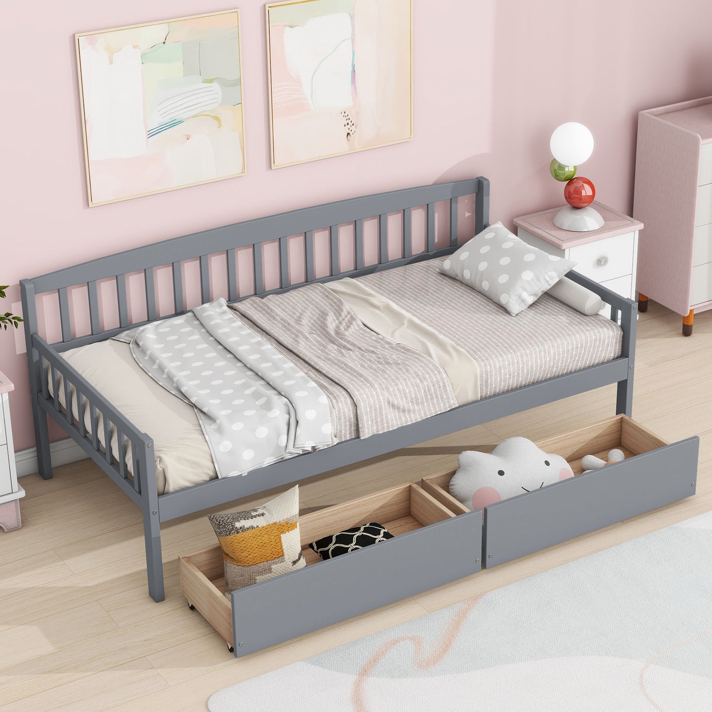 A Pine Wood Daybed with two storage drawers and a platform of 10 support slats sits against a pink wall, topped with polka-dot bedding and pillows. Plush toys fill the drawers, and a nightstand plus framed art complete the cozy setup.