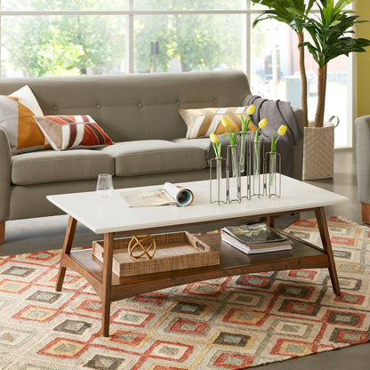 A modern living room features a gray sofa, a colorful patterned rug, and the Modern Table With Extra Storage holding a glass, magazine, and metal vase with yellow tulips. Sunlight pours in through large windows behind the scene.