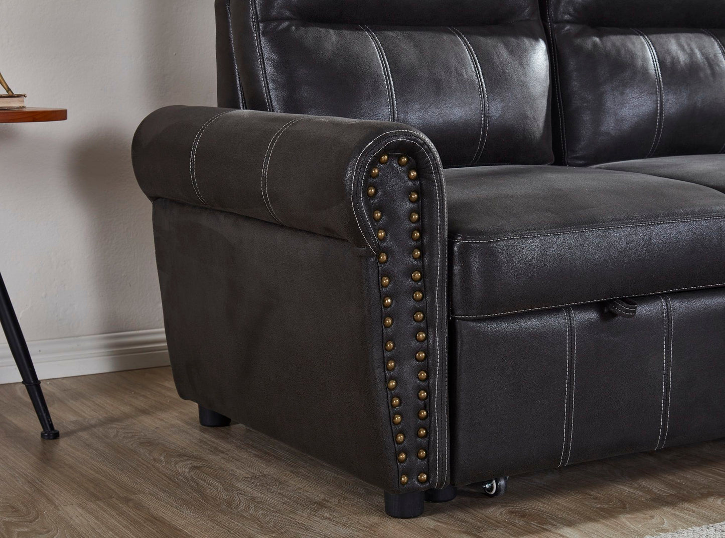 A close-up of the Comfort Sectional Sofa Bed features dark leather upholstery, rolled armrest, decorative brass nailhead trim, visible stitching, and wood flooring underneath.
