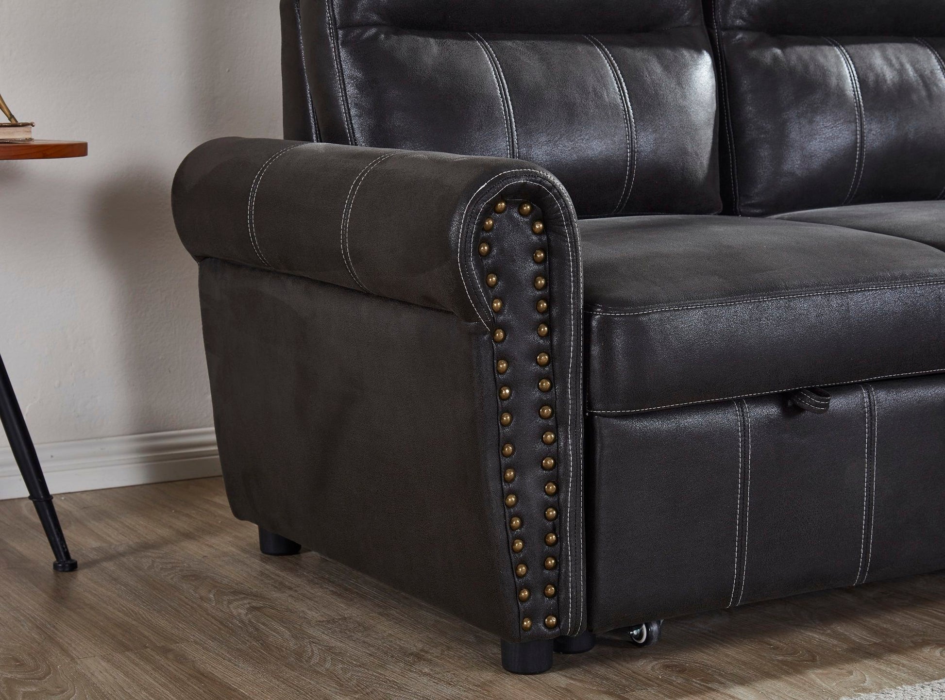 A close-up of the Comfort Sectional Sofa Bed features dark leather upholstery, rolled armrest, decorative brass nailhead trim, visible stitching, and wood flooring underneath.