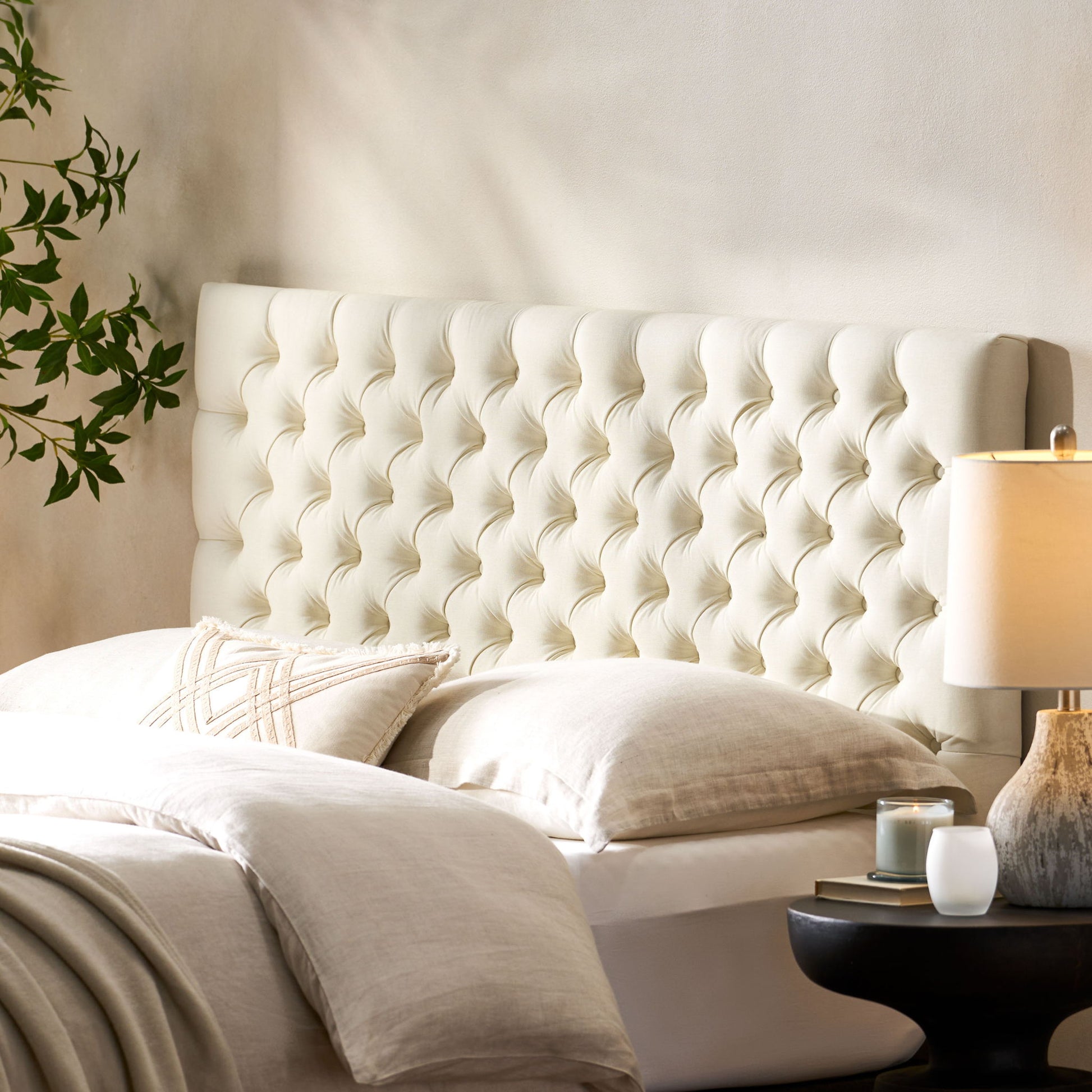 A cozy bedroom features the Annecy Contemporary Upholstered Headboard in cream, beige bedding with a textured pillow, a bedside table with a lamp and candle, and a leafy plant in the background.