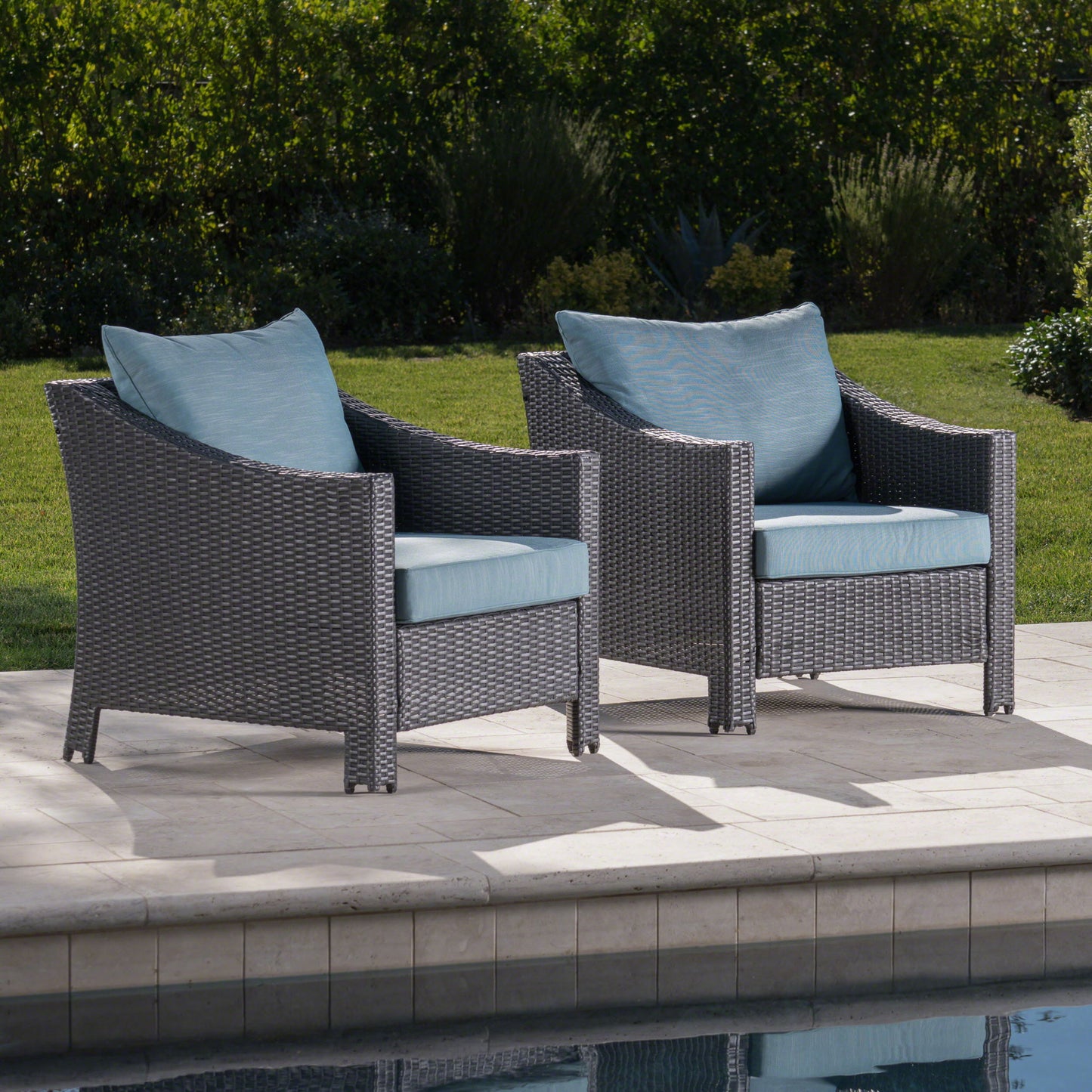 Two Antibes Armchairs with water-resistant blue cushions and pillows are set on a poolside patio, surrounded by green grass and shrubs.