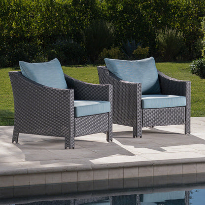 Two Antibes Armchairs with water-resistant blue cushions and pillows are set on a poolside patio, surrounded by green grass and shrubs.