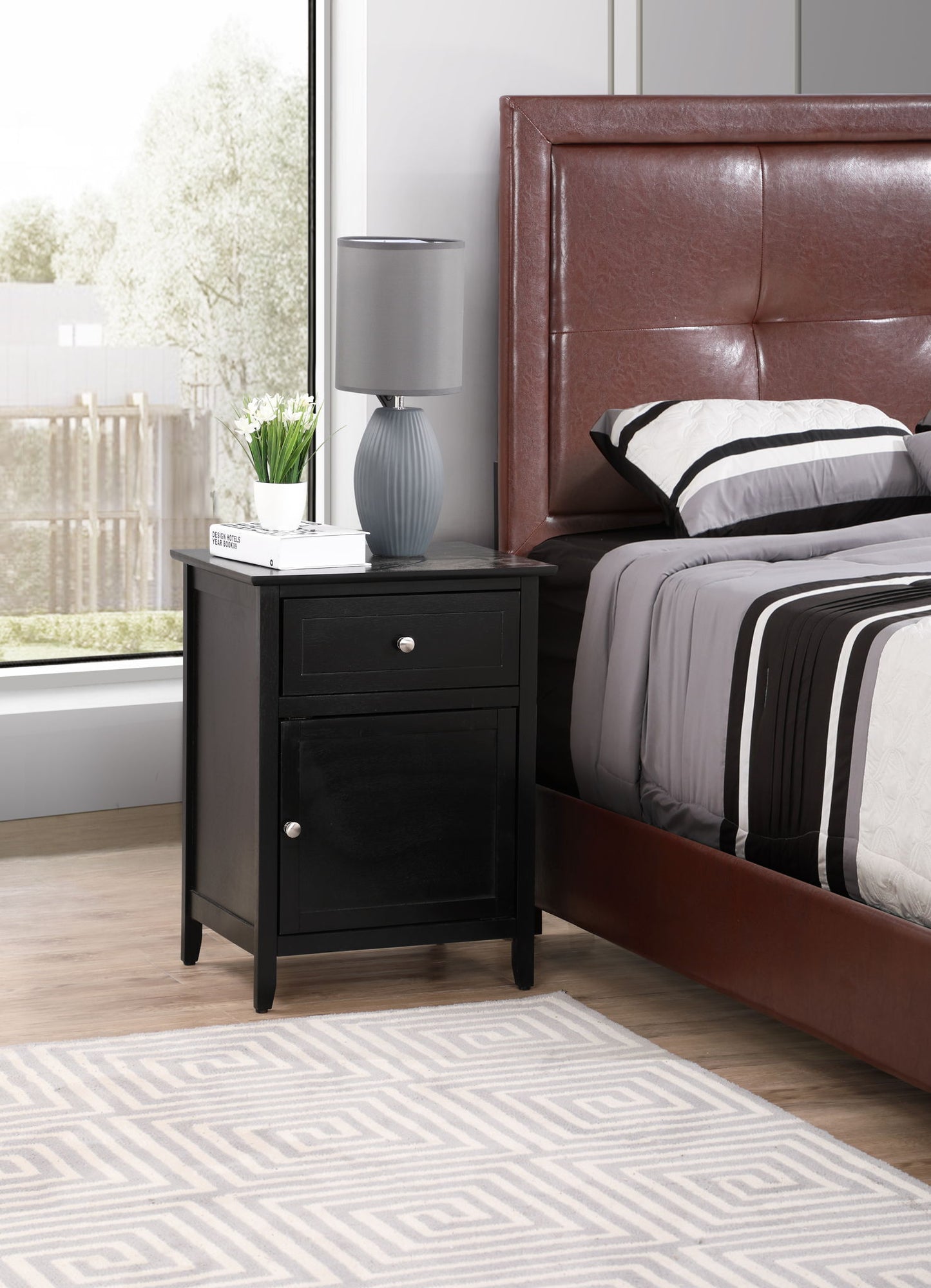 A modern bedroom with a brown upholstered bed, gray and white bedding, the Izzy 1 Drawer/1 Door Nightstand holding a lamp, potted plant, and book, beside a large window with trees outside.