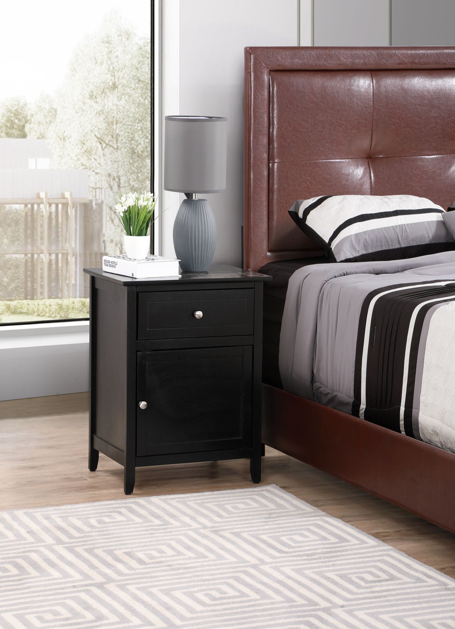 A modern bedroom with a brown upholstered bed, gray and white bedding, the Izzy 1 Drawer/1 Door Nightstand holding a lamp, potted plant, and book, beside a large window with trees outside.