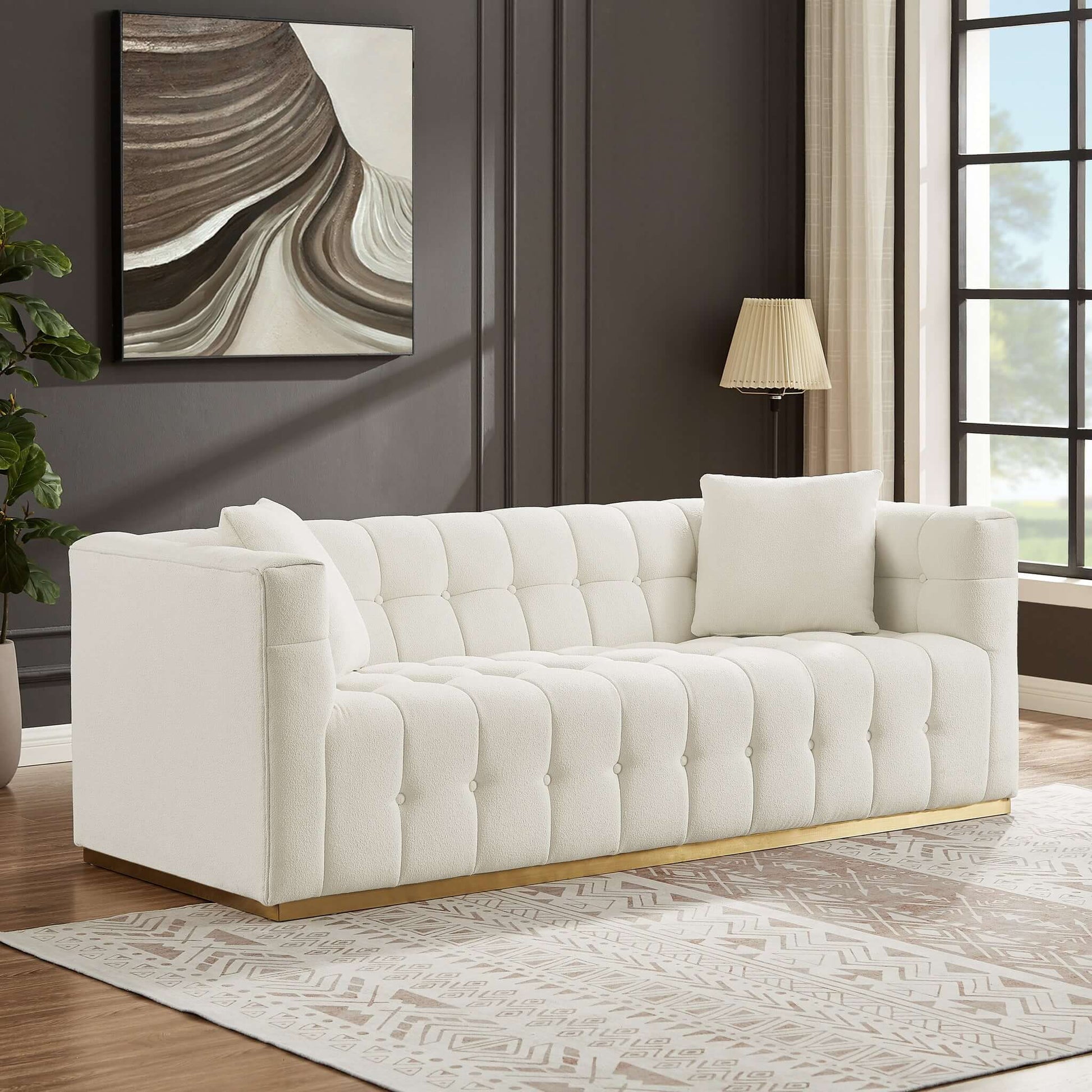 A modern living room showcases an Eleanor Sofa Boucle in ivory white with two cushions, a geometric-patterned rug, and a gold floor lamp. A large abstract painting adorns the dark wall as sunlight pours through expansive windows.