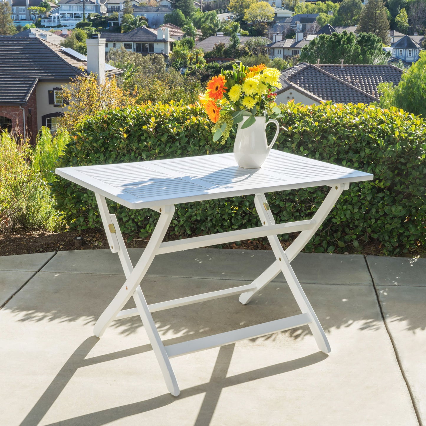 The Blissique Outdoor Foldable Wood Dining Table with a slat design is set on a patio, decorated with a vibrant floral pitcher, backed by green hedges and houses under a sunny sky.