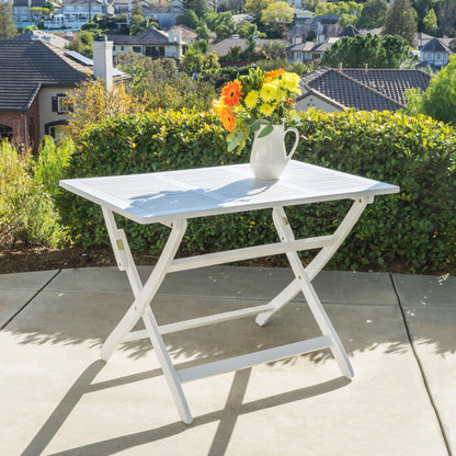 The Blissique Outdoor Foldable Wood Dining Table with a slat design is set on a patio, decorated with a vibrant floral pitcher, backed by green hedges and houses under a sunny sky.
